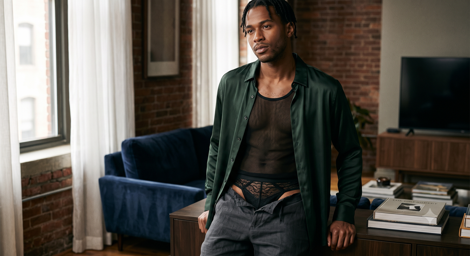 A male model showcasing men's lingerie, wearing a black mesh top and black lace briefs styled under an open emerald green satin shirt in a modern loft setting.